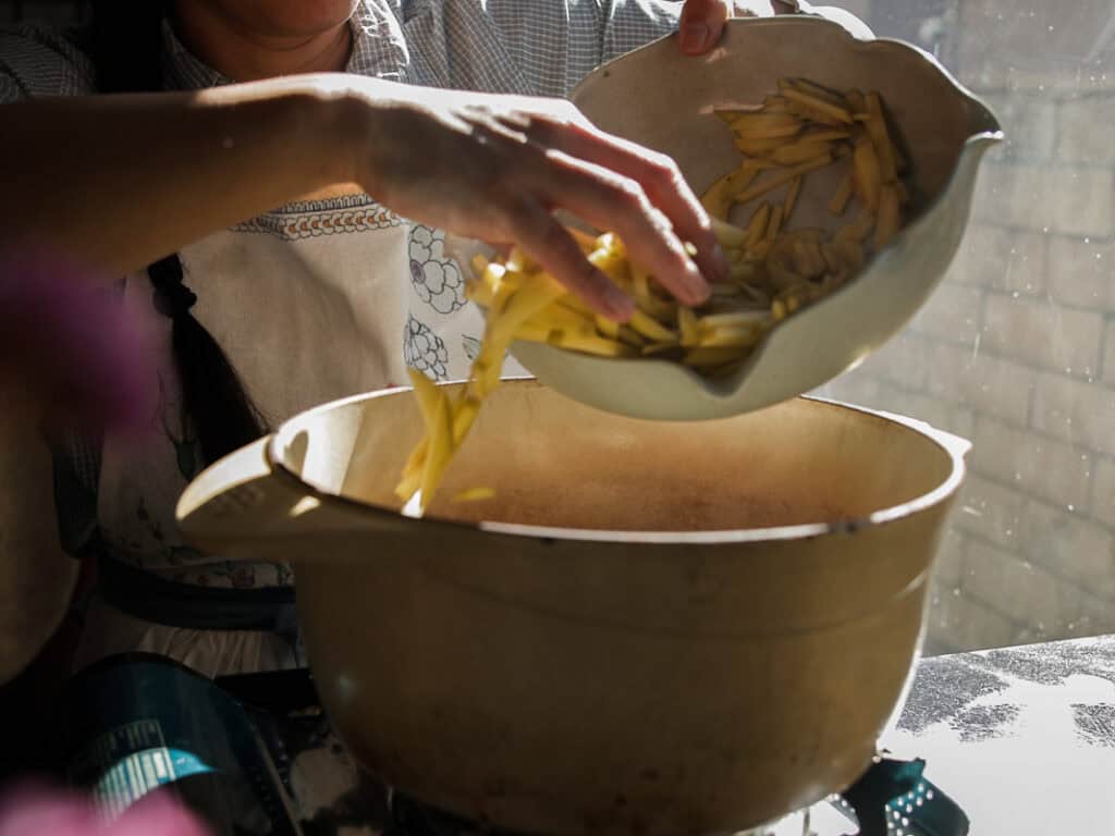 A person wearing an apron is adding sliced potatoes from a bowl into a large pot. Sunlight is streaming through a window, casting a warm glow over the scene.